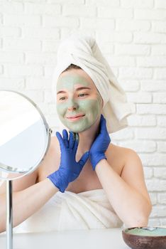 Spa and wellness. Natural cosmetics. Self care. Happy young caucasian woman in white bath towels wearing gloves applying clay face mask looking at the mirror