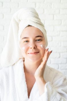 Spa and wellness. Natural cosmetics. Self care. Happy young caucasian woman in white bath towel applying face cream at home