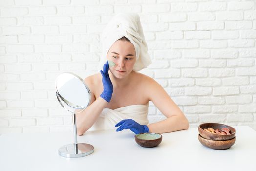 Spa and wellness. Natural cosmetics. Self care. Close up of happy young caucasian woman in white bath towels wearing gloves applying clay face mask looking at the mirror