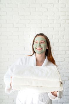 Spa and wellness. Natural cosmetics. Self care. Young smiling caucasian woman with a face mask wearing bathrobes holding a pile of white bath towels