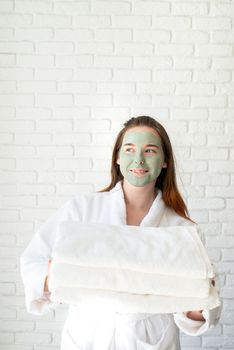 Spa and wellness. Natural cosmetics. Self care. Young smiling caucasian woman with a face mask wearing bathrobes holding a pile of white bath towels