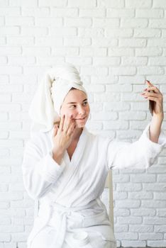 Spa and wellness. Natural cosmetics. Self care. Happy young caucasian woman in white bath towel applying face cream at home doing selfie