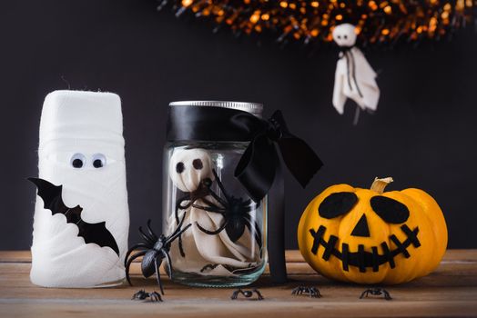 Funny Halloween day decoration party, White ghost scary face and spider in jar glass, glass of water wrapped with bandage and pumpkin on wood table, studio shot isolated on black background