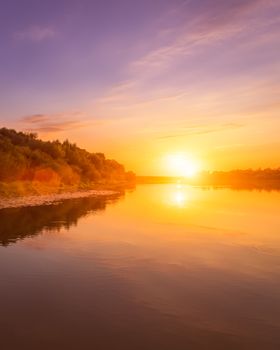 Sunset or dawn on a river with a cloudy sky and sun rays reflected in the water, water lilies and trees growing along the shore. Landscape.