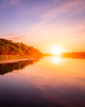 Sunset or dawn on a river with a cloudy sky and sun rays reflected in the water, water lilies and trees growing along the shore. Landscape.