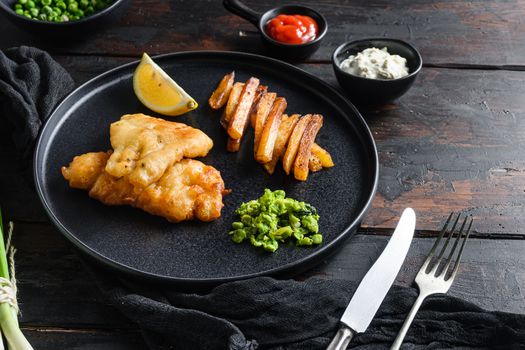 Fish & chips with dip and lemon black plate with British traditional cusine food with beer-battered cod and fries and a side of tartar sauce mushy minty peas served in the Pub or Restaurant space for text side view.
