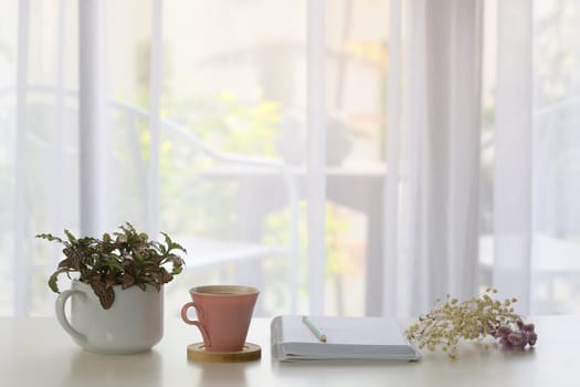 Interior house decor with pink coffee cup and white notebook and blue pencil and plants with see through curtain