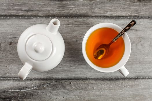 Table top view on small white teapot and cup full of hot amber tea with silver spoon in it, placed on gray wood table.