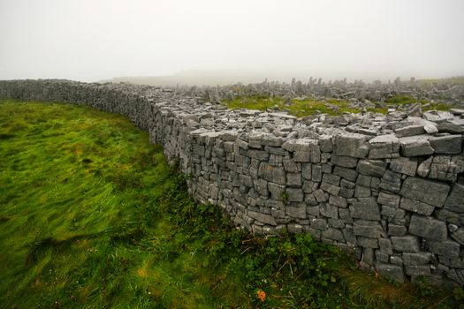 Stone brick walls rising from soft grass near Dun Aonghasa - semi circular stone fort on Inis Mor (Inishmore) island with thick fog in background. Aran Islands, Ireland.