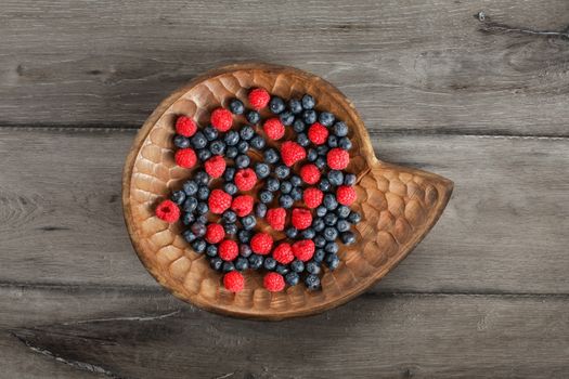 Table top view on mixed blueberries and raspberries in vintage carved wooden plate placed on gray wood desk.