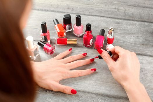 View from behind to a woman hand, applying second coat of red nail varnish on her nails, with lot of nail polish on gray wood desk in background.