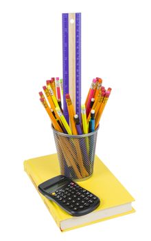 Vertical shot of pencils and a ruler in pencil holder with calculator and pencil holder resting on a brightly colored yellow book isolated on white. 