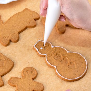 Young woman is decorating Christmas Gingerbread House cookies biscuit at home with frosting topping in icing bag, close up, lifestyle.