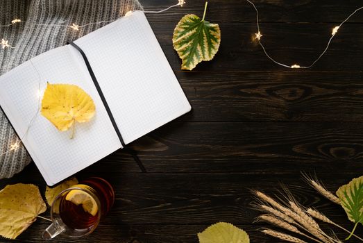 Autumn composition. Workspace with notebook, cup of lemon tea, autumn leaves and fairy lights. Top view, flat lay on black wooden background