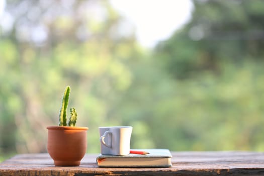 White cup with notebook and brown clay cactus pot on wooden table with nature view