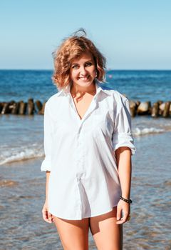 young blonde smiling woman in a white swimsuit and shirt posing standing on the seashore on sunny summer day