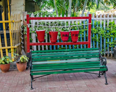 Freshly painted, old classic fire extinguishing water buckets & old wooden bench with cast frame on a railway platform in India.
