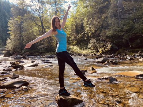 young woman doing yoga exercises on the stone among mountain stream