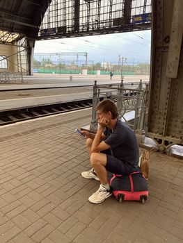 young man sitting on a bag on the railway station and watching on the mobile phone