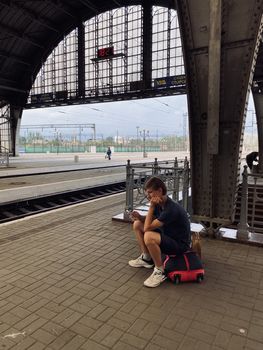 young man sitting on a bag on the railway station and watching on the mobile phone