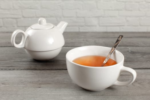 White ceramic teapot and cup of hot amber tea on gray wood desk.