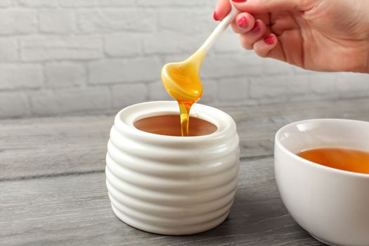 Detail on woman holding small ceramic  spoon, filled with honey, about to put it in white tea cup full of hot tea.