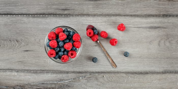 Table top view on small glass bowl filled with mix of blueberries and raspberries, silver spoon on the side, placed on gray wood desk.