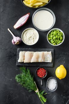 Organic fish and chips ingredients raw cod fillets on stone slate batter, potatoe, tartar sauce, minty mushy peas, lemon , shallot, mint, garlic, salt, peppercorns on black stone background overhead photo vertical.