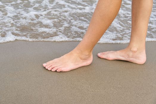 Close-up of child's legs walking on sand coast and sea water. Young girl legs on baltic sea beach. Teenager legs near sea line