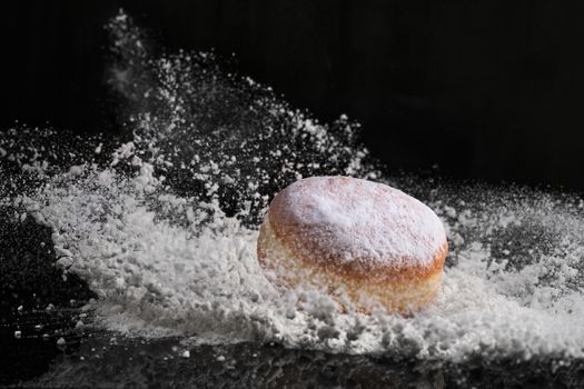 Berliner donut with jam stuffed falls into flour on black background.