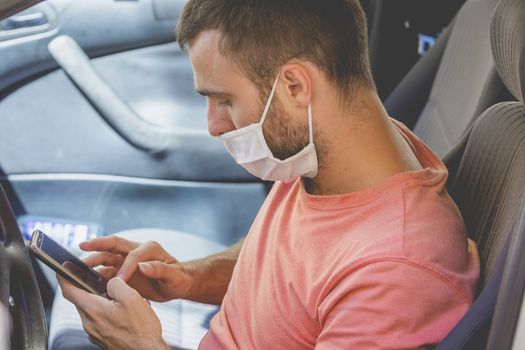 A young man, wearing a cloth face mask, checks the mobile phone inside the car, while he waits for someone before drive, in the province of Zaragoza, Spain.