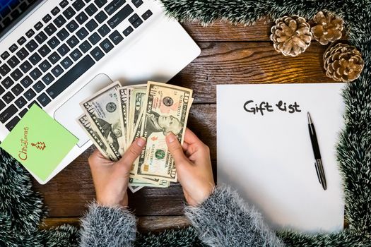 Hand holding money for buying Christmas gifts. Empty gift list on decorated office table. Top view of Christmas composition .