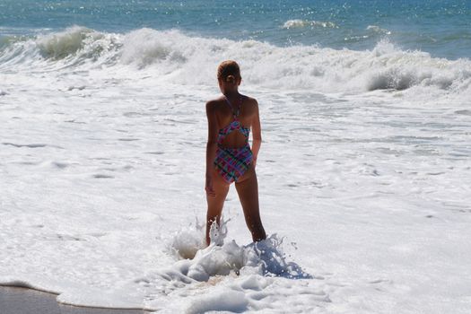 teenage girl standing on the beach in sea white foam, back view
