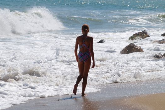 teenage girl walking alone along the empty seashore on the sunny day, rear view