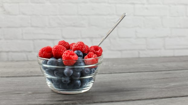 Small glass bowl full of blueberries with raspberries on top, and silver spoon, placed on gray wooden desk with white brick wall in background.