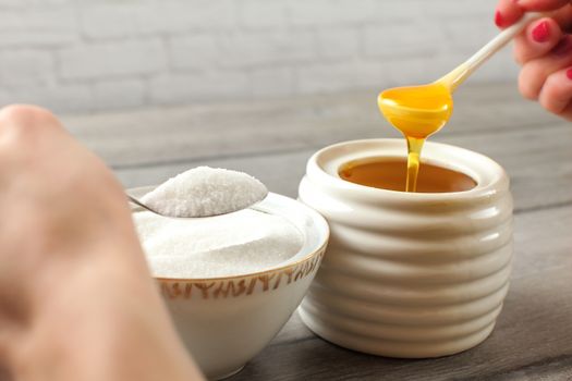 Detail on woman hand, one holding honey dipper with natural honey, other silver spoon with refined white sugar. Sugar vs honey concept.