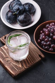 Fresh ice cold carbonated water in glass on cutting board with rosemary leaf near to wooden bowls with grape and plum fruits, dark stone background, angle view