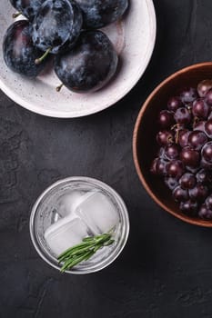 Fresh ice cold carbonated water in glass with rosemary leaf near to wooden bowls with grape and plum fruits, dark stone background, top view