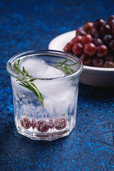 Fresh ice cold carbonated water in glass with rosemary leaf near to wooden bowl with grape berries, blue textured background, angle view macro