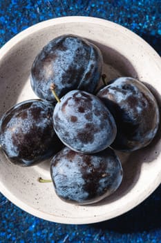 Fresh ripe plum fruits in white wooden bowl on blue abstract background, top view