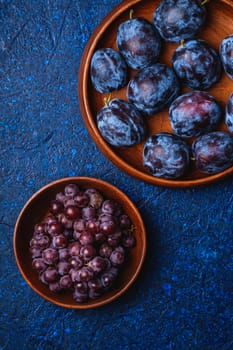 Fresh ripe plum fruits in brown wooden plate and grape berries in bowl on blue abstract background, top view