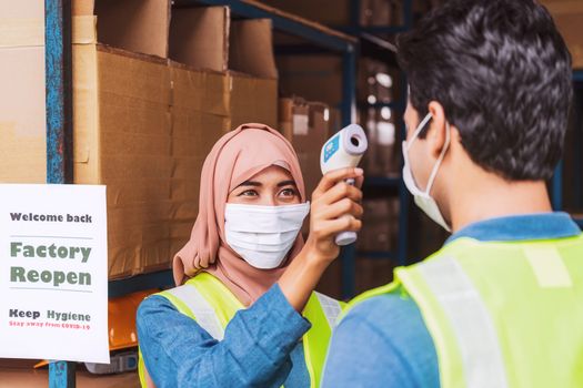 Muslim woman worker wearing surgical mask and Hijab uses Medical Digital Infrared Thermometer measure temperature to Indian man worker with safety clothes before start to work after warehouse reopen