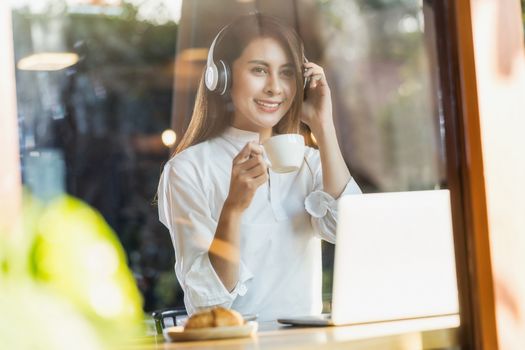 Portrait Young Asian woman holding a cup of coffee and using technology laptop in a coffee shop for working and listening music, freelancer and entrepreneur working by connecting to internet.