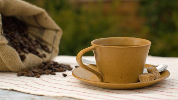 Close-up of coffee beans in a burlap jute sack and cup of coffee with raw sugar on a table in the garden