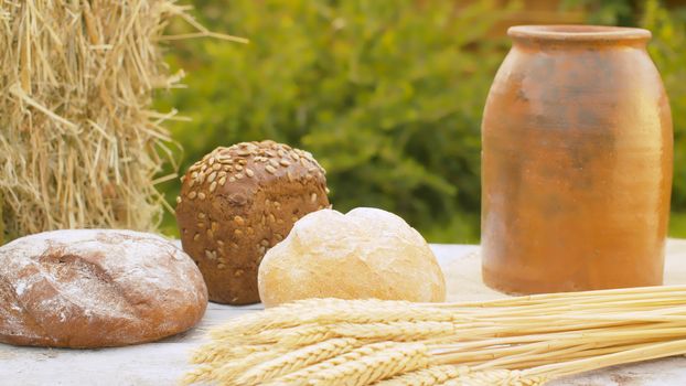 Close up loafs of traditional baked bread, ripe wheat ears, fresh hay and clay pot on table outdoors. Natural organic handmade food. Rural scene. Close-up shot with slide or move camera