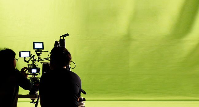Behind the video production camera set and crew team shooting in a big green screen studio.