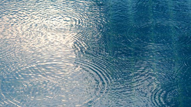 Rain drops on the swimming pool blue water surface that have ripple wave effect on liquid texture and top view angle.