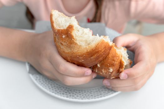 Pretty little girl eats a fresh croissant for breakfast. Portrait of cute girl with yummy face enjoy eating croissant. Healthy food for children concept.