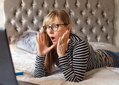 woman in glasses lying on a bed looking at a laptop and wondering