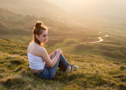A girl sitting on top of the hill against amazing landscape at sunset in the summer